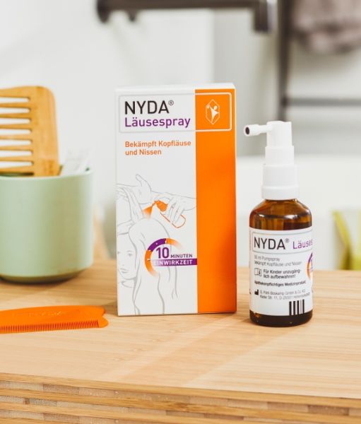 NYDA® Läusespray Flasche und Verpackung auf einem Holztisch, daneben ein orangefarbener Läusekamm, im Hintergrund Badezimmerutensilien.