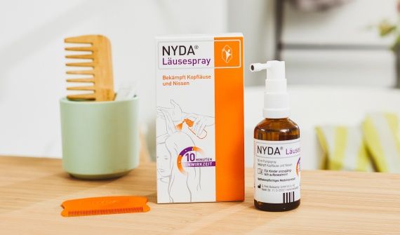 NYDA® Läusespray mit Verpackung, Flasche und orangefarbenem Läusekamm auf einem Holztisch, im Hintergrund ein Holzkamm in einem Becher.
