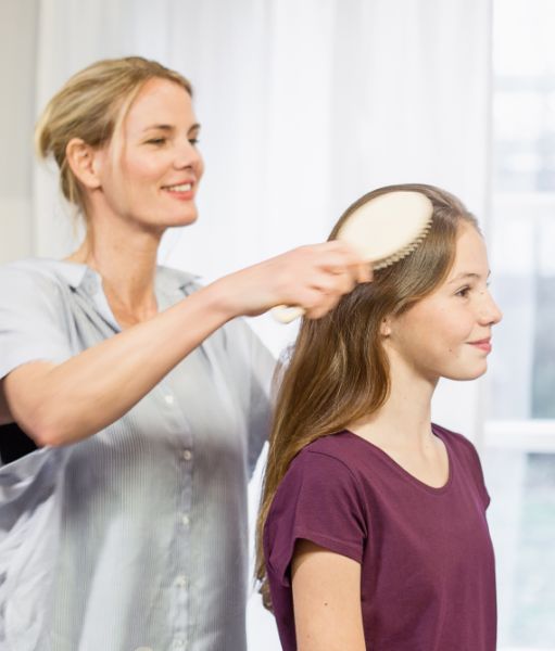 Frau bürstet das Haar eines Mädchens mit einer Haarkurzbürste in einem hellen Raum.
