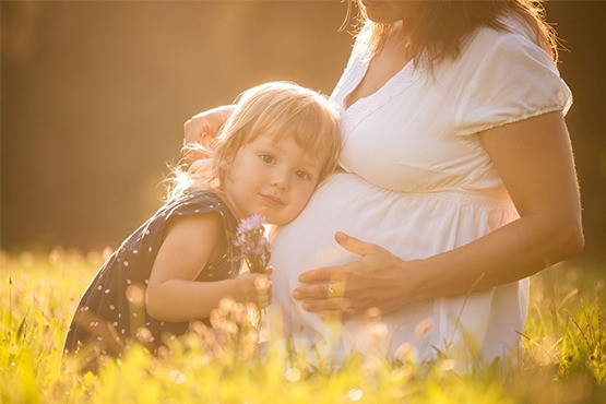 Ein kleines Mädchen lehnt sich sanft an den Bauch einer schwangeren Frau und hält einen Blumenstrauß. Beide sitzen in einem sonnigen Feld, die Szene wirkt warm und friedlich.