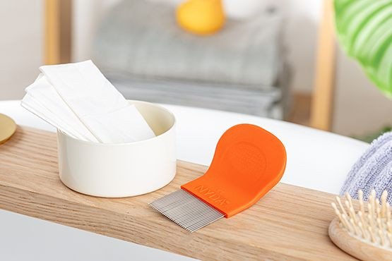 Orangefarbener NYDA® Läuse- und Nissenkamm liegt neben einem Becher auf einer Holzablage, umgeben von weiteren Pflegeutensilien.