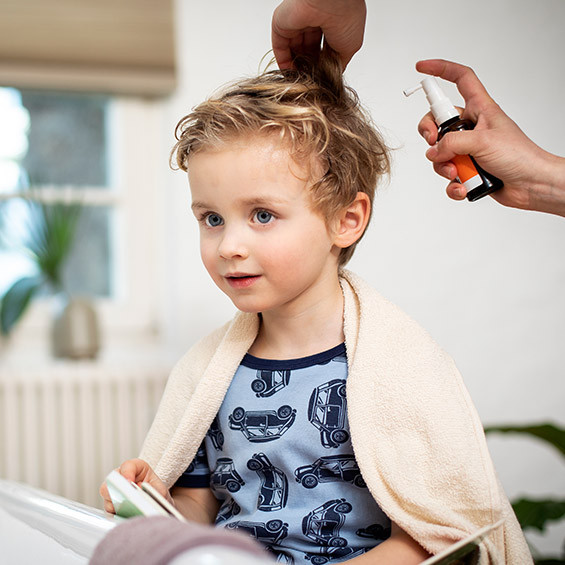 Ein kleines Kind mit blonden, leicht lockigen Haaren sitzt ruhig, während eine Person ein Läusespray auf sein Haar aufträgt. Das Kind trägt ein Handtuch über den Schultern.