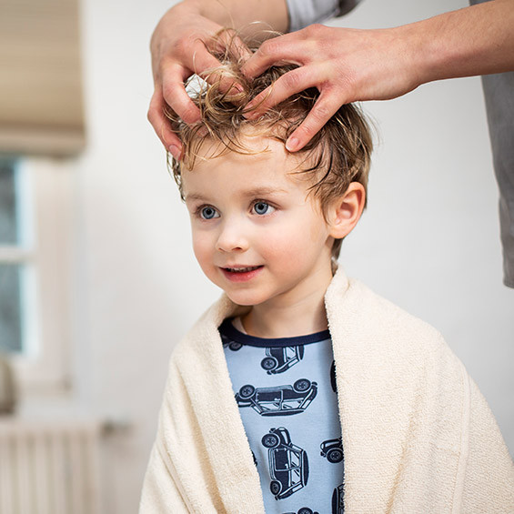 in kleines Kind mit blonden Haaren lächelt, während eine Person die Flüssigkeit in sein Haar einmassiert. Das Kind trägt ein Handtuch über den Schultern und ein Hemd mit Autoprint.