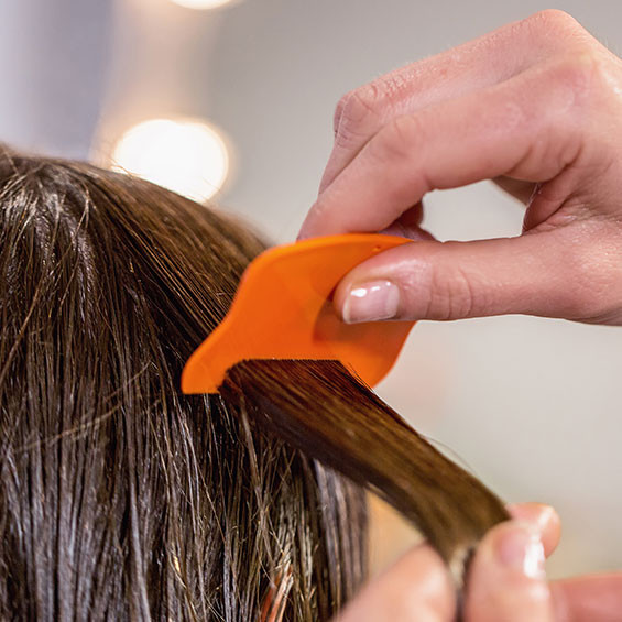 Entfernen von abgetöteten Läusen und Nissen mit dem NYDA® Läusekamm. Eine Nahaufnahme zeigt eine Hand, die mit einem orangefarbenen Läusekamm nasse Haarsträhnen durchkämmt, um abgetötete Läuse und Nissen zu entfernen.