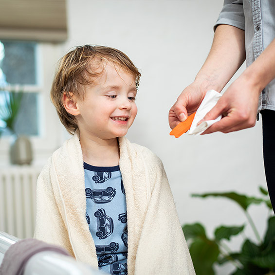 Ein kleines Kind mit blonden, nassen Haaren sitzt mit einem Handtuch um die Schultern und lächelt. Eine Person hält einen orangefarbenen Läusekamm und reinigt ihn mit einem Papiertuch.