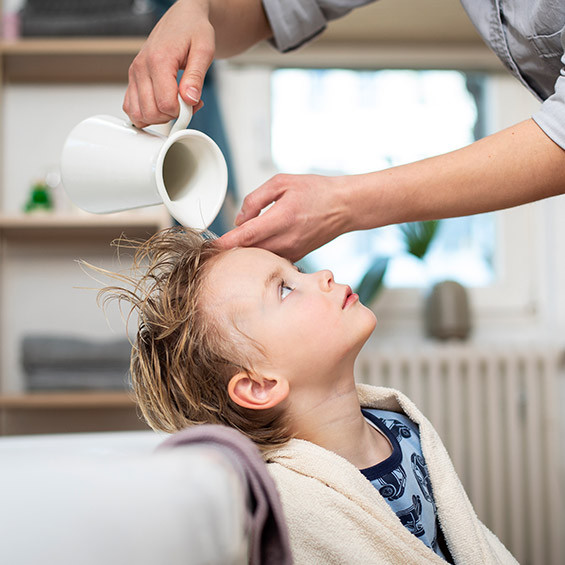 Ein kleines Kind mit blonden, nassen Haaren lehnt sich zurück, während eine Person mit einer Kanne Wasser über das Haar gießt. Das Kind trägt ein Handtuch über den Schultern und sitzt im Badezimmer.