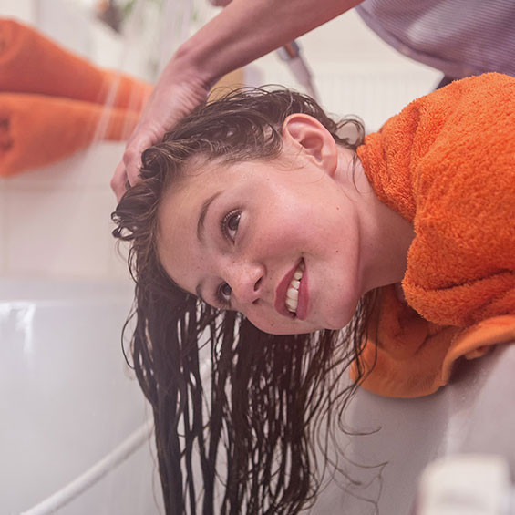 Nach der NYDA® Anwendung die Haare gründlich mit einem normalen Shampoo auswaschen. Ein Mädchen mit nassen Haaren beugt sich über die Badewanne, während eine Person ihr die Haare mit Wasser ausspült. Sie trägt ein orangefarbenes Handtuch um die Schultern und lächelt.