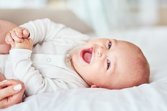 Lächelndes Baby in weißem Strampler liegt auf dem Bett und hält die Hände zusammen.