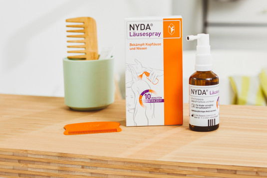 NYDA Läusespray mit Pipette und Kamm auf Holzunterlage neben grünem Behälter.